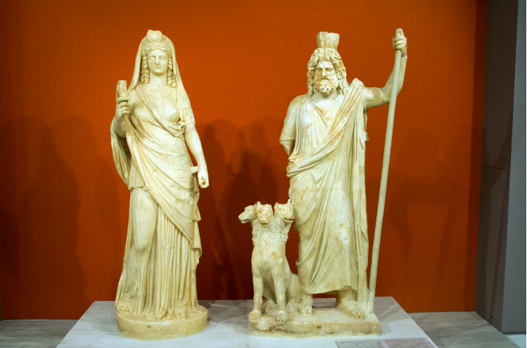 two roman statues, woman on the left and man on the right, wearing a jar on his head, a sceptre and with a dog