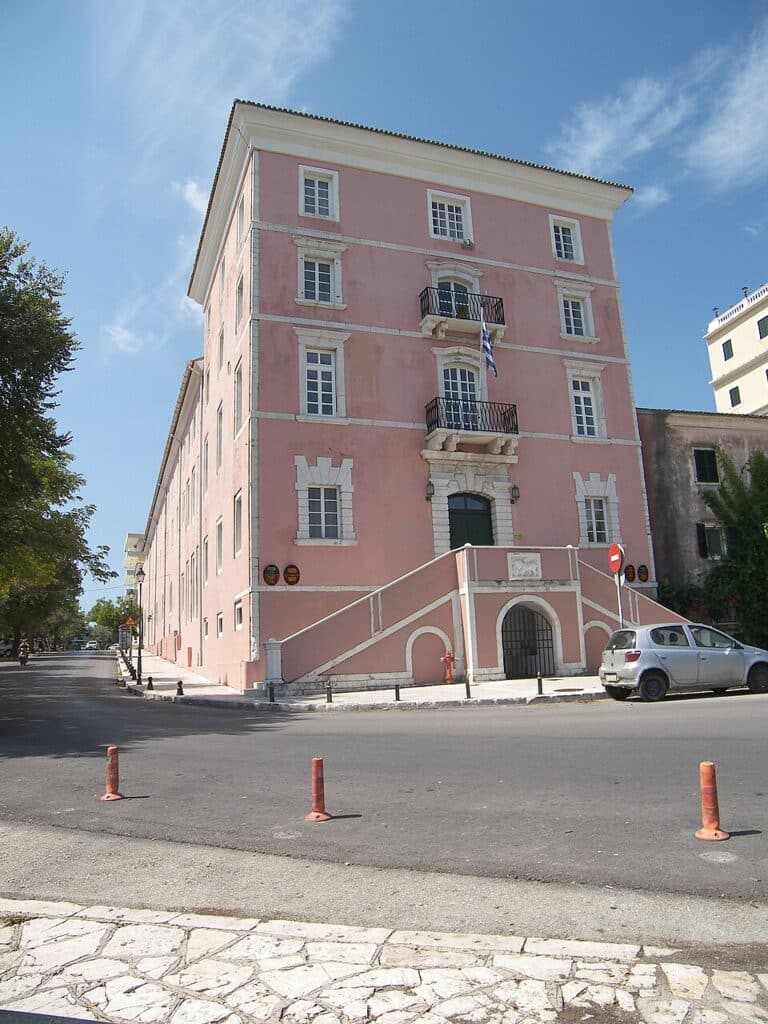 A tall, pink three-story building with a distinctive double staircase and white stone trim sits at a quiet street intersection on Corfu.