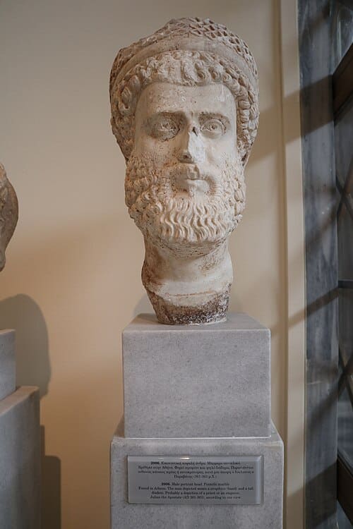 A sculpted marble portrait head of a bearded man wearing a diadem rests upon a stone pedestal inside a museum.
