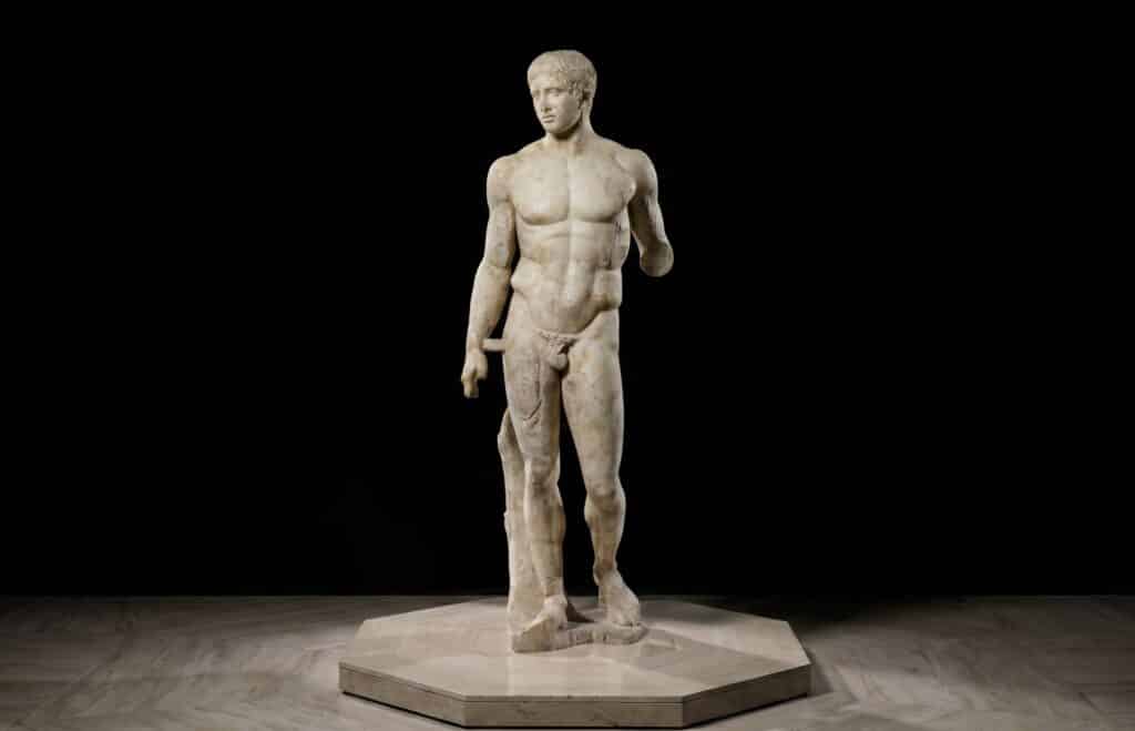 A white marble statue of a nude male figure, known as the Spear-Bearer or the Doryphoros, by Polykleitos, stands with one arm bent and the other by his side on a stone base against a black background.