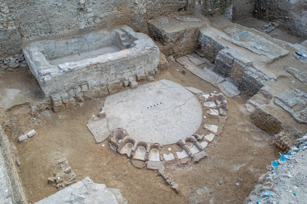 An overhead view of an archaeological excavation reveals the stone foundations of a circular structure with individual seating niches alongside a rectangular basin.