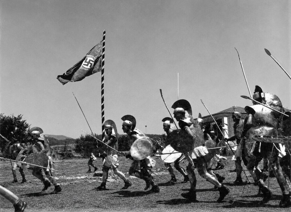 Several men wearing replica ancient Greek armor charge across a grassy field carrying spears and shields, with a large Nazi swastika flag flying prominently in the background.