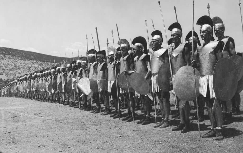 A long line of men dressed in replica ancient Greek hoplite armor and modern military boots stand in formation holding spears and shields in a dry, mountainous landscape.