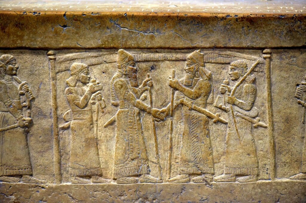 An ancient carved stone relief depicts two central figures in ornate robes shaking hands, flanked by attendants holding staffs and parasols.