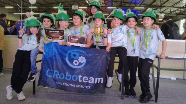 Greek elementary school students from the GRoboVines robotics team pose with awards, medals, and a team banner at a robotics competition.