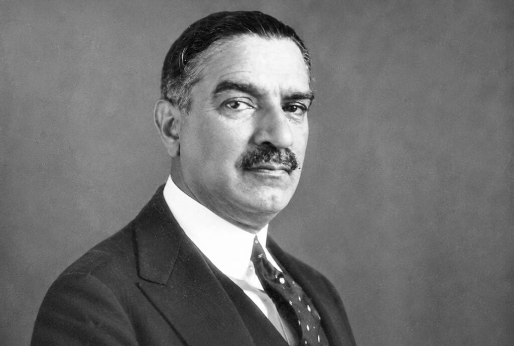 A black-and-white portrait features Greek politician Andreas Michalakopoulos wearing a dark suit, white collared shirt, and a patterned tie.