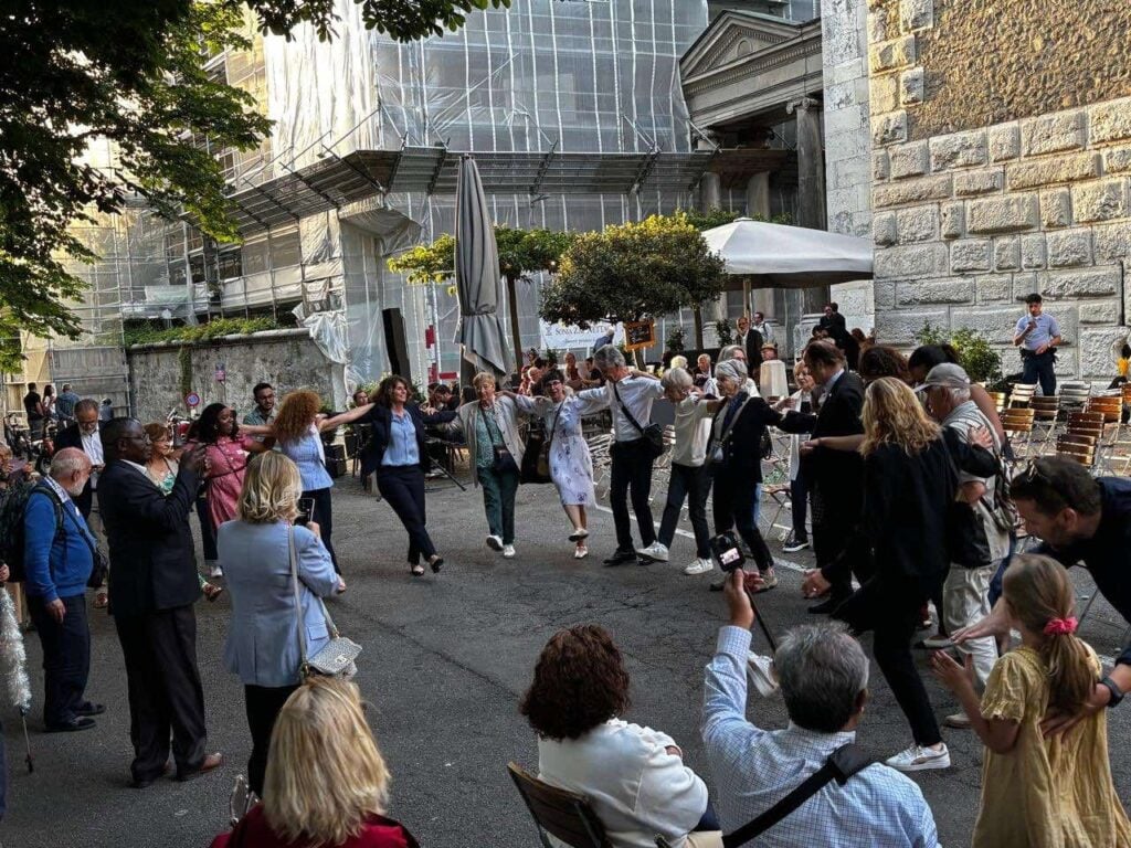 A large group of people holds hands in a circle to perform a traditional Greek dance on a paved outdoor square while spectators look on and take photos.