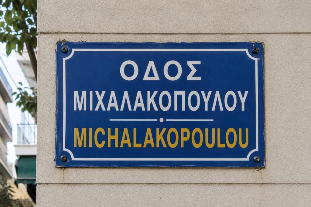 A blue enameled street sign mounted on a textured beige wall displays the name "Michalakopoulou" in white Greek lettering and yellow Latin characters.