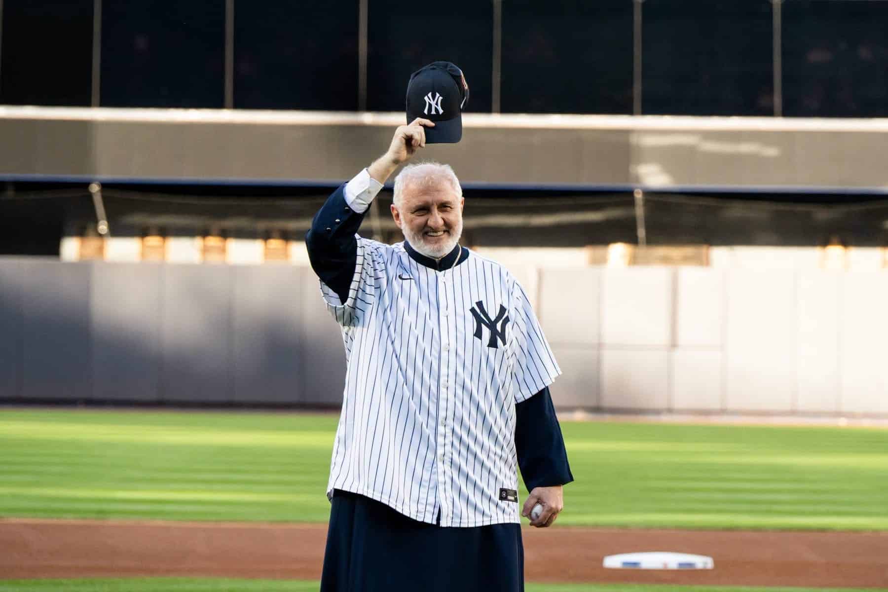 Archbishop Elpidophoros wearing a Yankees jersey and raising a Yankees cap at Yankee Stadium during Greek Jersey Night.