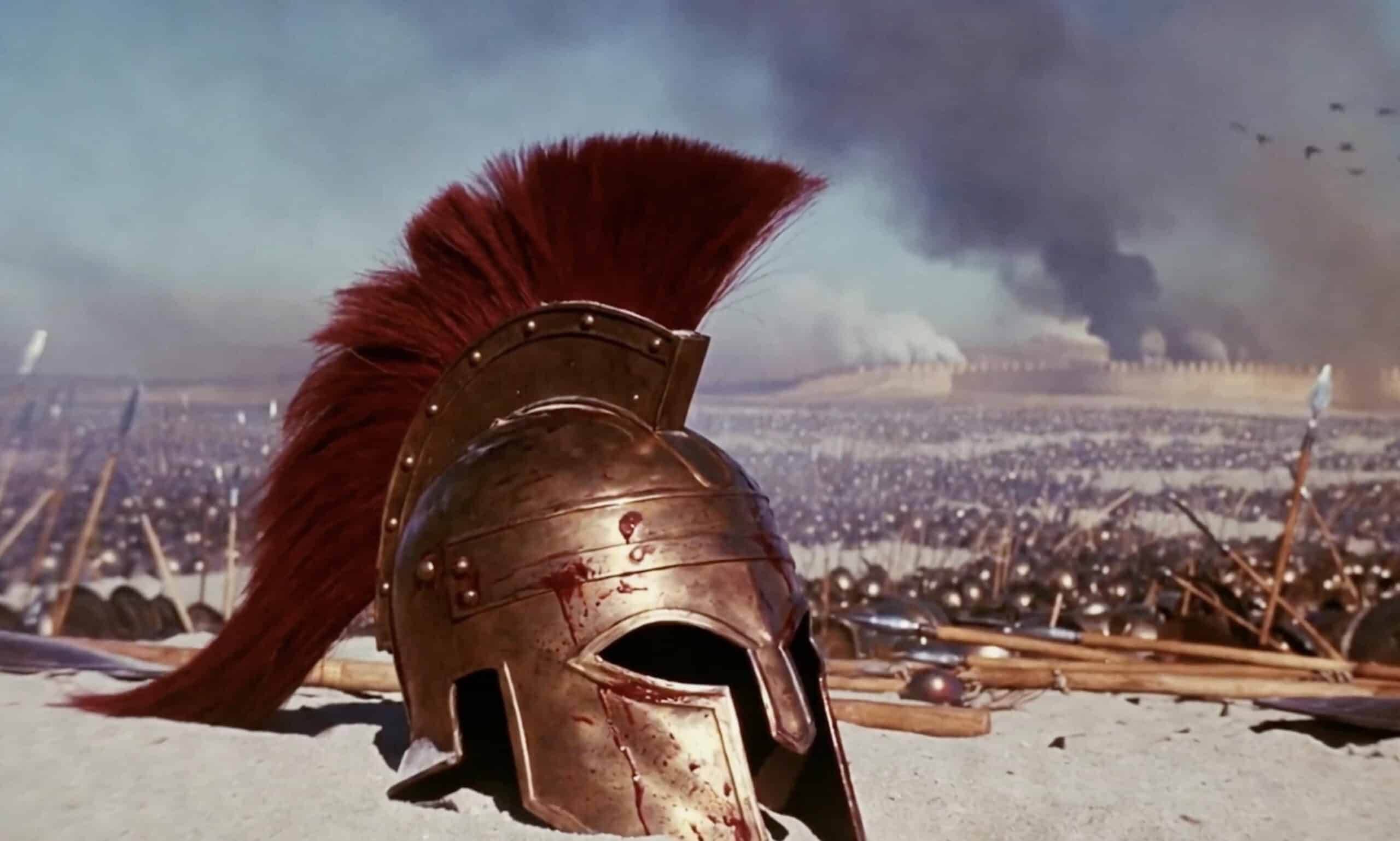 A bronze ancient Greek-style warrior helmet with a red plume lies on a battlefield, with spears, shields, smoke, and armies in the background.