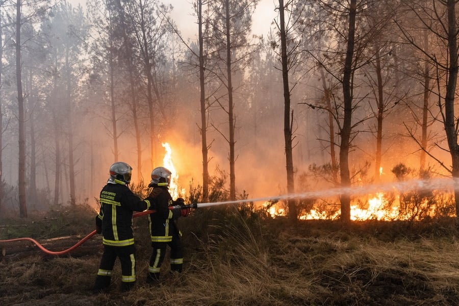 Two firefighters spray water on flames spreading through a smoke-filled forest during a major wildfire in France.