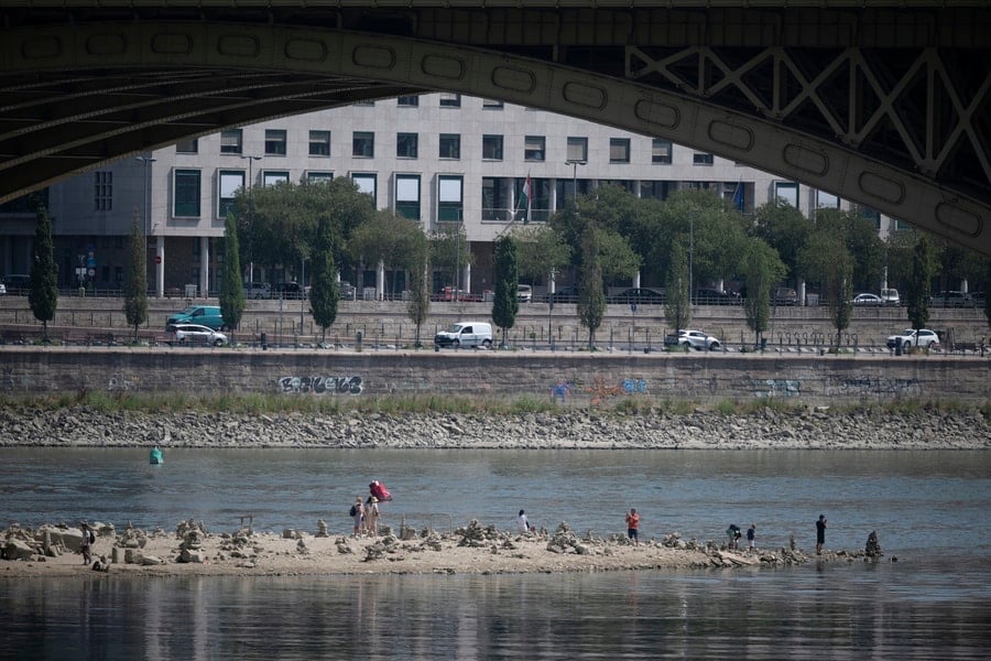 People stand on an exposed section of the Danube riverbed as low water levels reveal rocks and sand beneath a bridge.