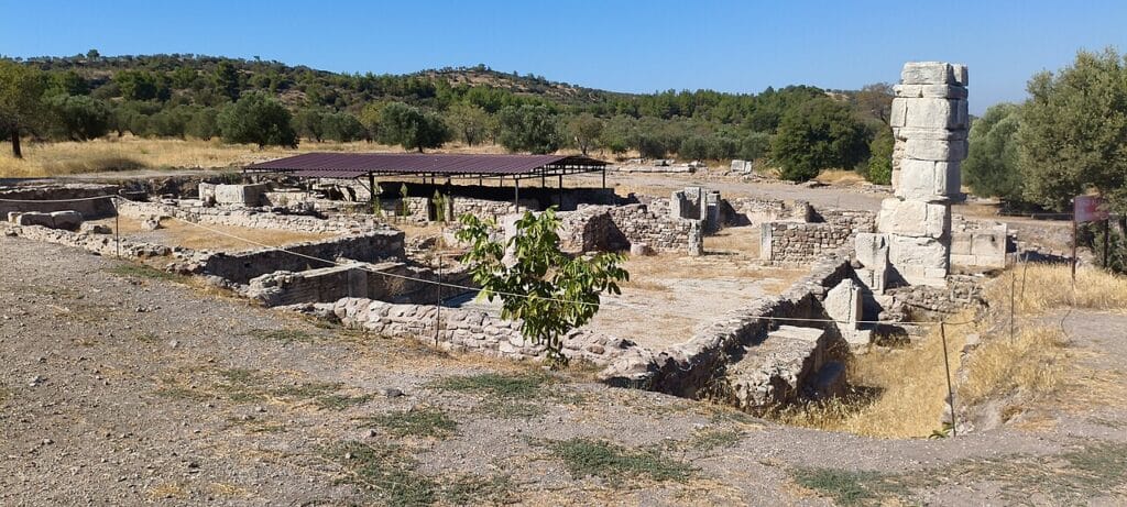 A sunlit view of an archaeological site features exposed stone foundations, a partial stone column, and a modern protective awning set amidst a dry landscape with rolling hills.