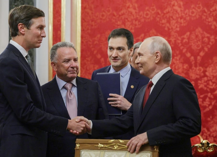 Russian President Vladimir Putin shakes hands with a tall American delegate while two other men observe the greeting in a room decorated with red patterned walls.