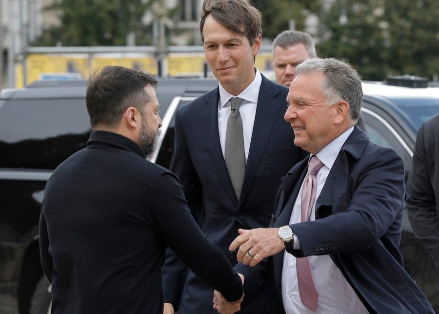 Ukrainian President Volodymyr Zelenskyy shakes hands with an older American delegate wearing a pink tie, as a younger delegate stands behind them smiling outdoors.