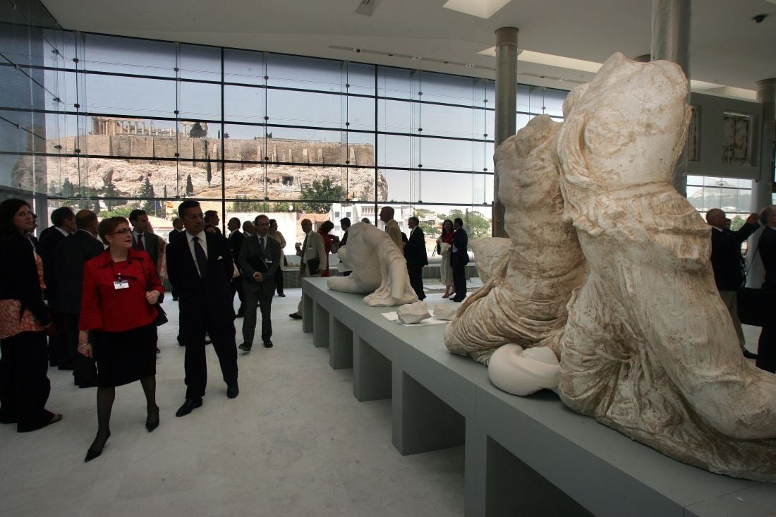Visitors explore the Acropolis Museum in Athens, with the Acropolis visible through the glass windows.
