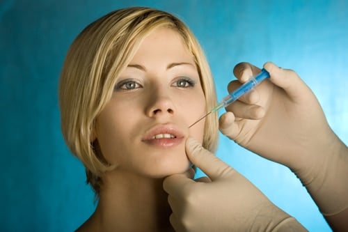 A close-up shot of a blonde woman receiving a cosmetic injection near her lips from a medical practitioner wearing white gloves.