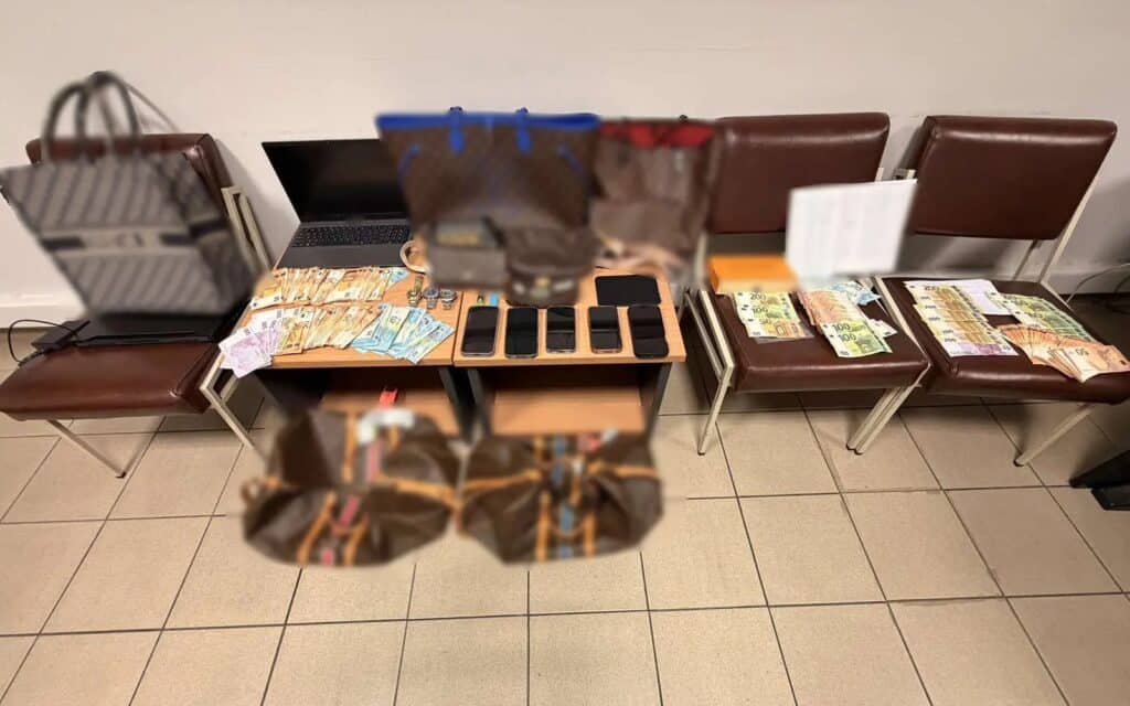 A display of confiscated items including blurred luxury handbags, a laptop, smartphones, and scattered Euro banknotes resting on four brown chairs.
