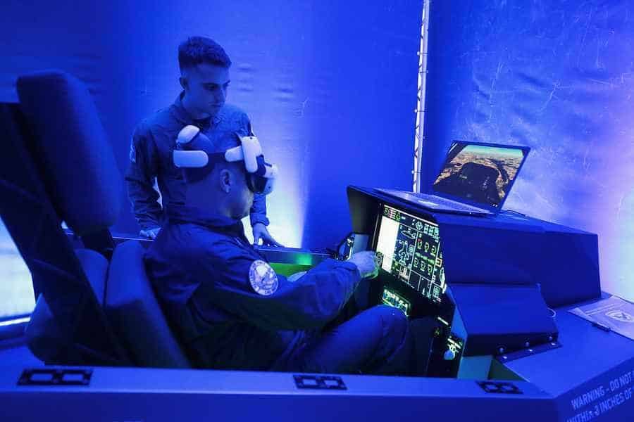 An instructor in military overalls monitors a visitor wearing a virtual reality headset inside a mockup fighter jet cockpit cockpit setup illuminated by bright blue lights.