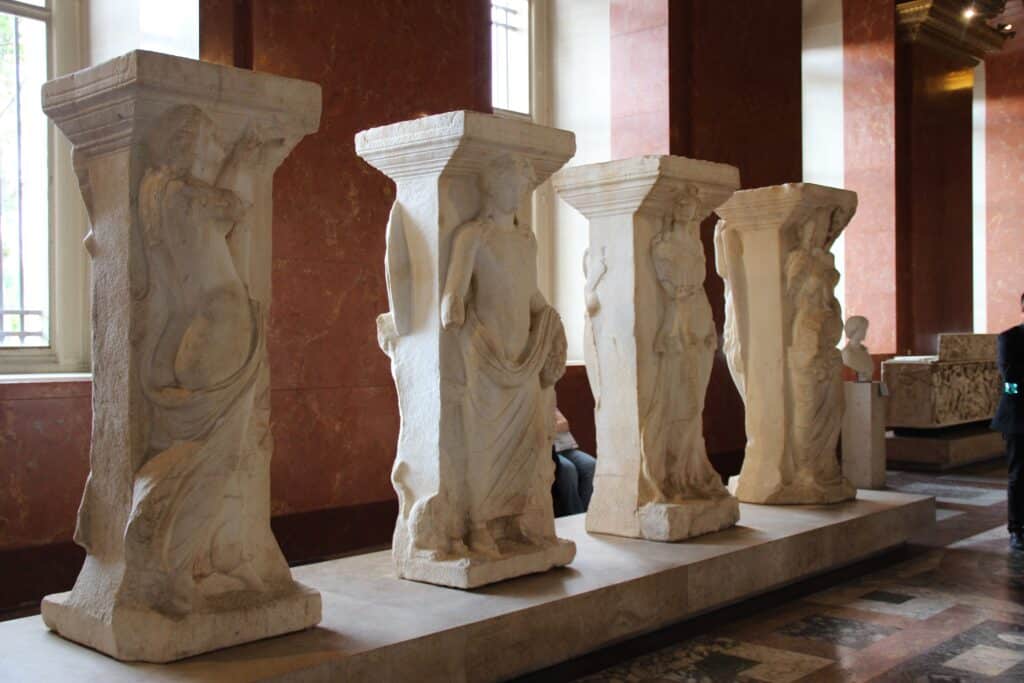Four double-sided Roman carved marble pillars with mythological figures standing in a row inside a Louvre Museum gallery.