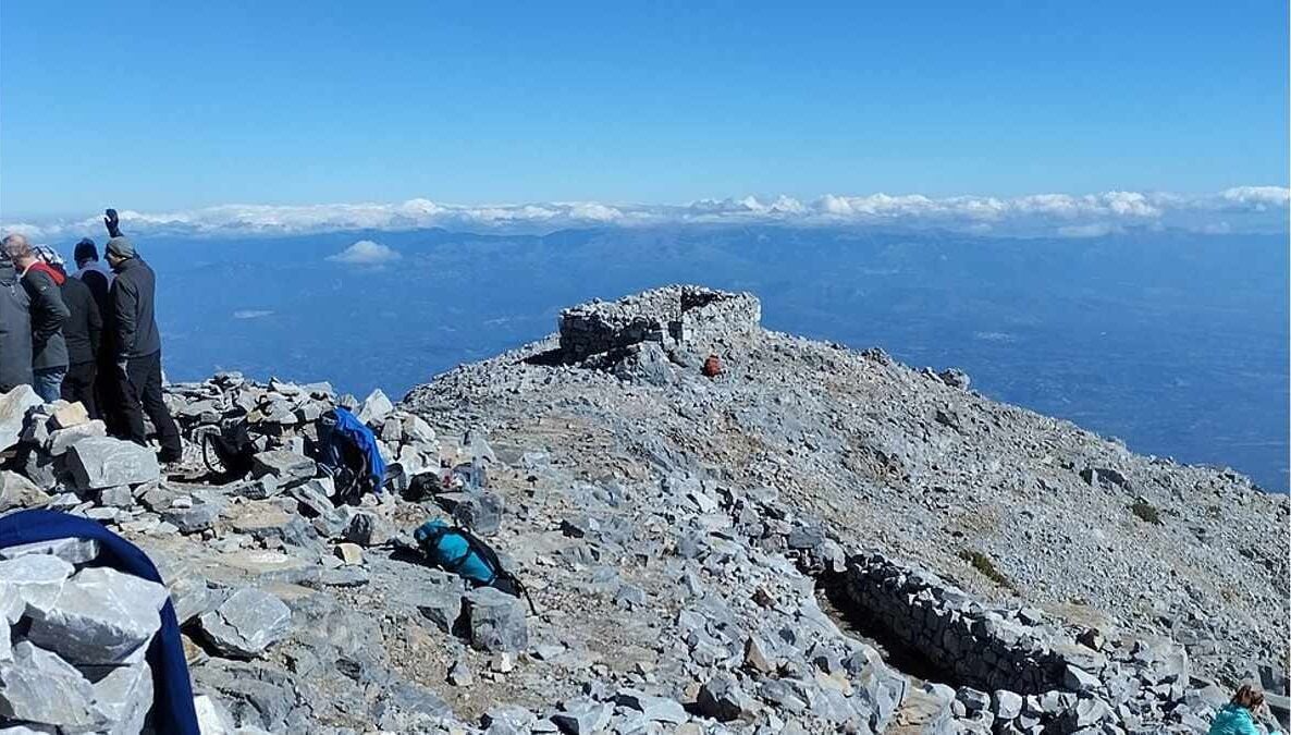 An expansive alpine panoramic photograph showcases multiple hikers gathering on the rocky, stone-covered summit of Mount Taygetos under a clear blue sky.