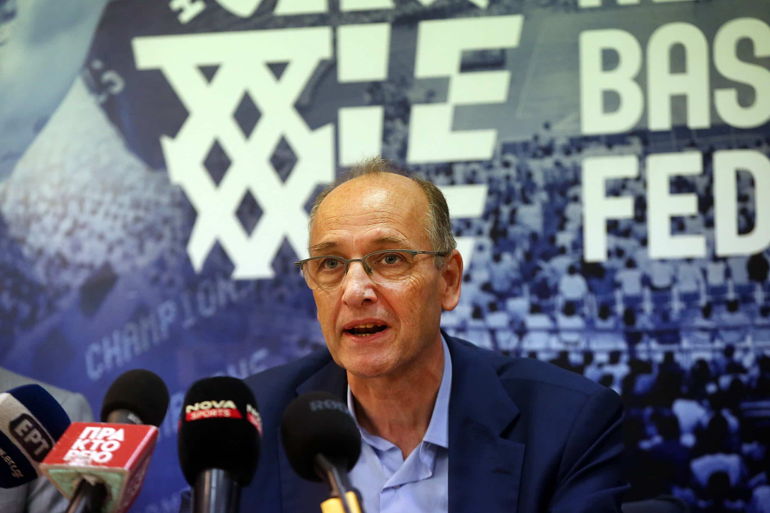Hellenic Basketball Federation President Vangelis Liolios and FIBA Secretary General Andreas Zagklis sit side by side in formal suits during an indoor sporting event.