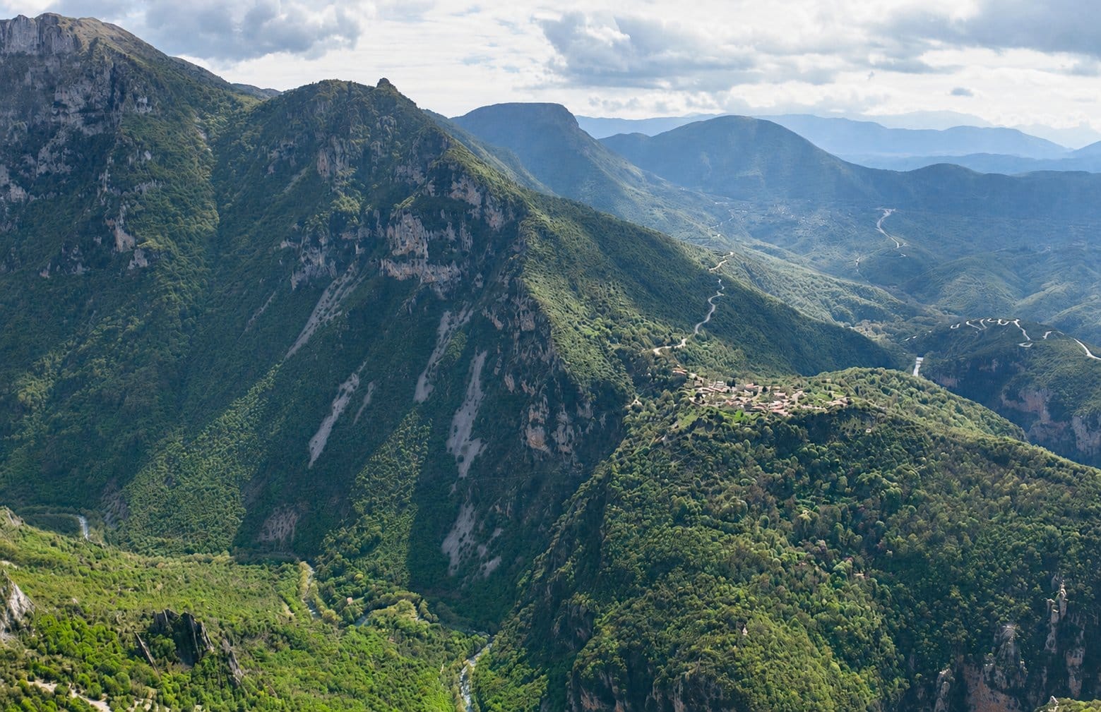 Vikos Gorge Pindus Greece : Forested mountain peaks surround a deep valley with a small hilltop village and winding roads.