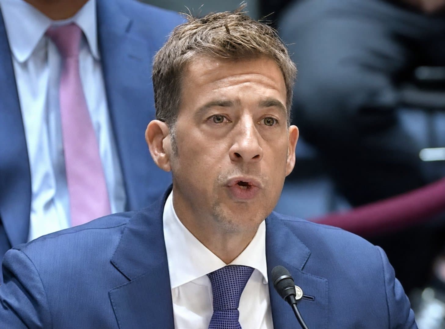 A man in a blue suit speaks into a microphone during a formal hearing.