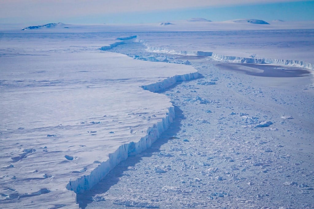 A vast Antarctic ice shelf fractured into jagged cliffs beside a channel filled with broken ice.