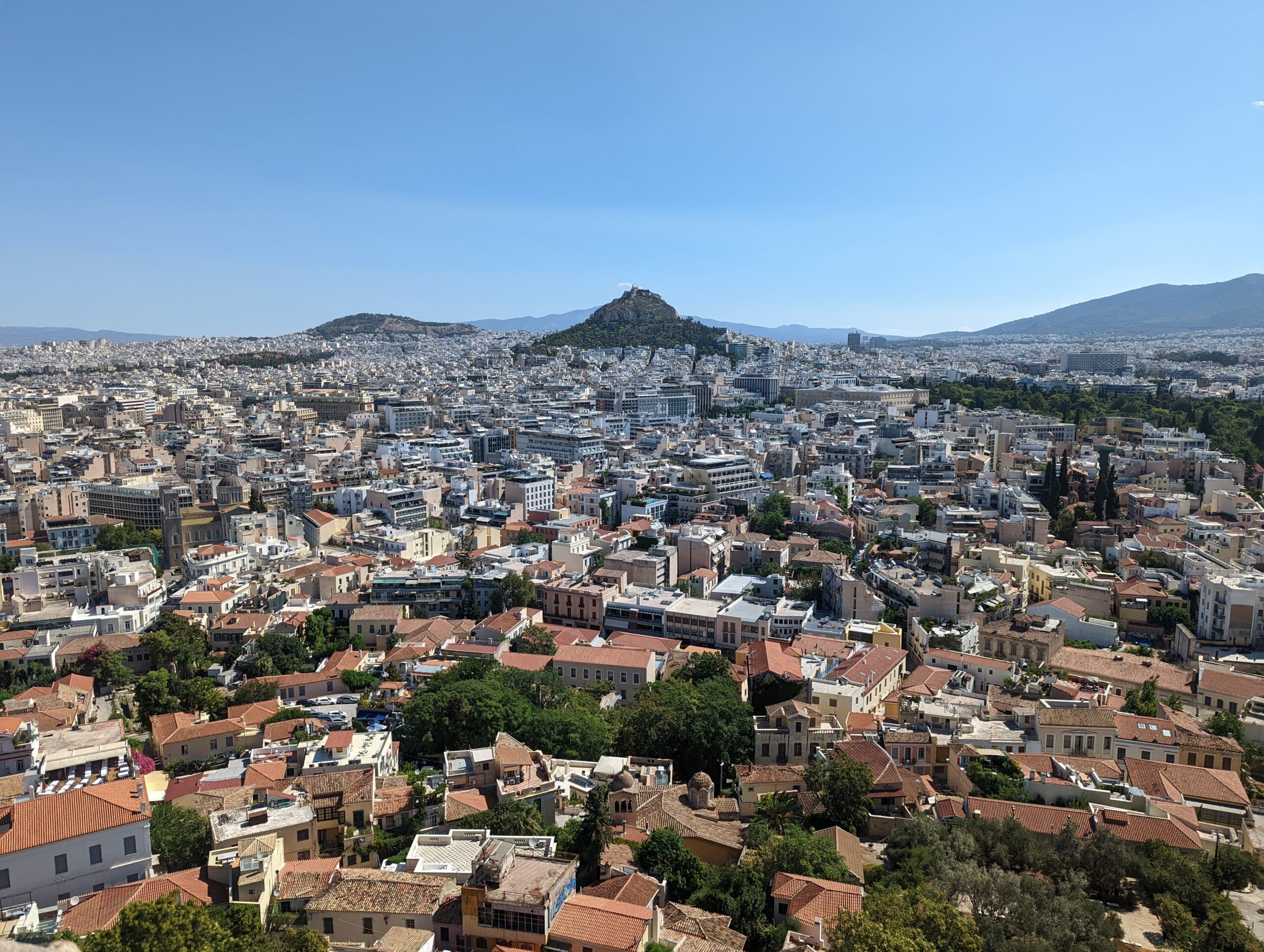 Dense buildings across Athens beneath a clear blue sky, with Lycabettus Hill in the distance.