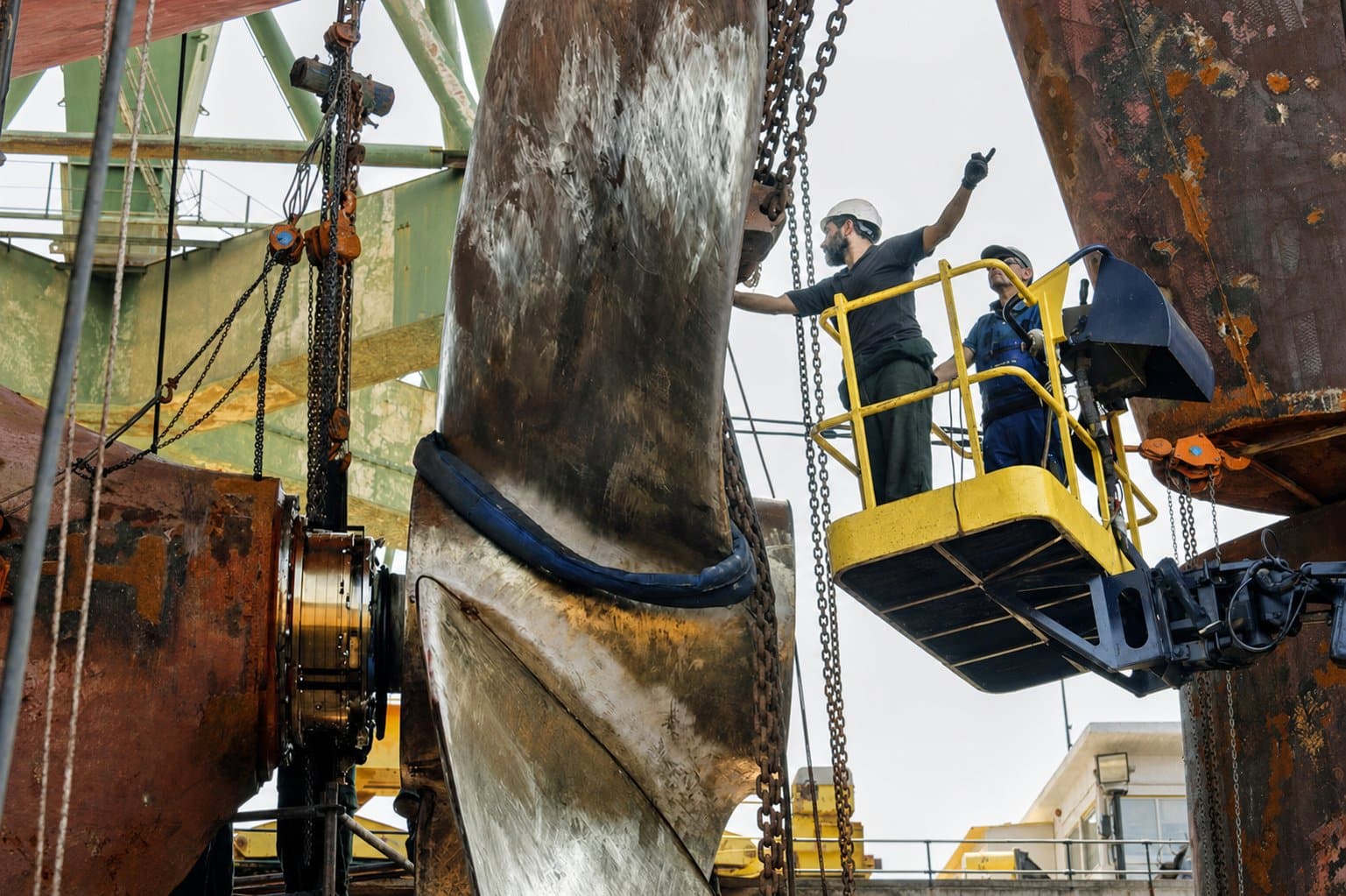 Two workers on a lift inspect a large ship propeller amid chains and industrial equipment.