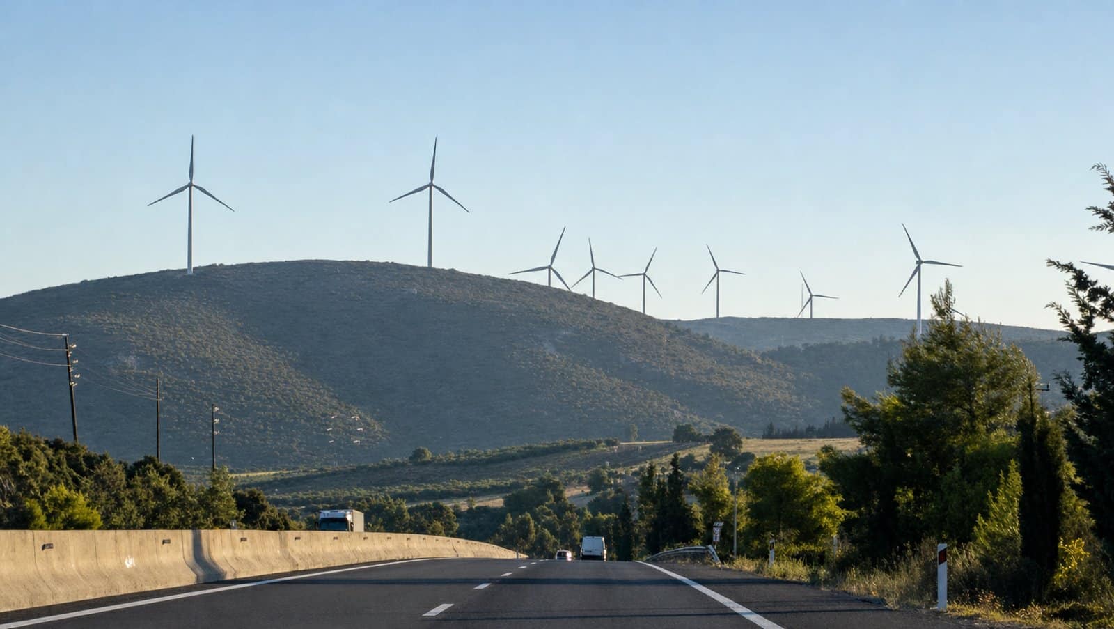 Wind turbines line sunlit hills above a highway bordered by trees.