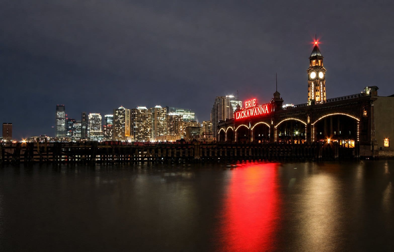 Illuminated Erie Lackawanna terminal and clock tower beside a nighttime city skyline across the water.