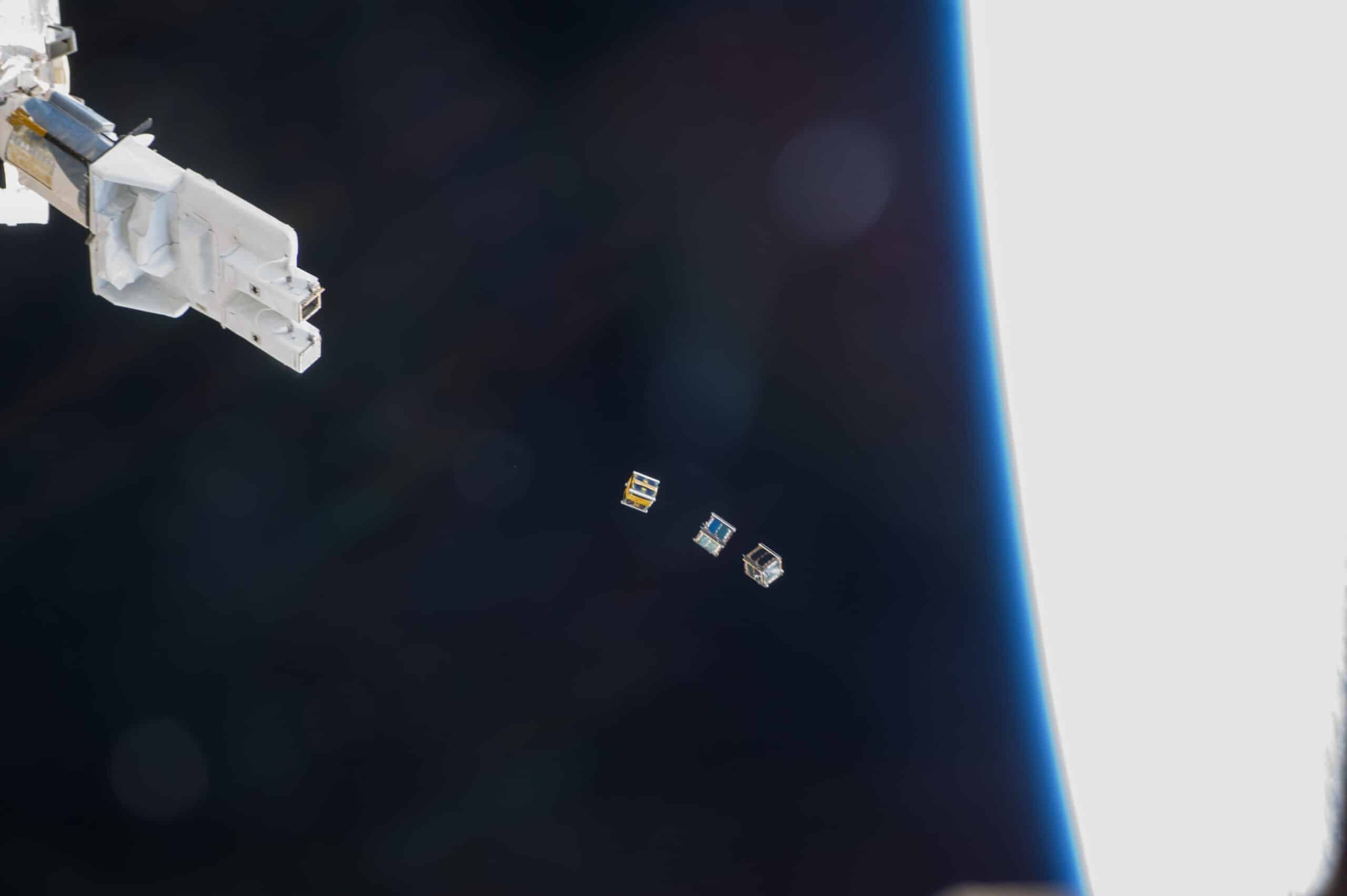 Three small CubeSats float near the International Space Station's robotic arm, with Earth behind them.